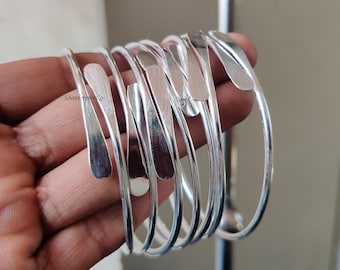 Puede incluir: Una colección de brazaletes de metal plateado. Cada brazalete tiene un diseño curvo y abierto con una pieza de acento plana y alargada. Los brazaletes están apilados, mostrando sus superficies reflectantes. La marca "SilverEmpireCo" es visible.