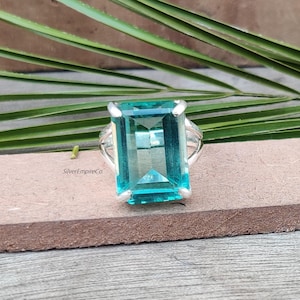 May include: A silver ring featuring a large, rectangular, light blue gemstone. The gemstone is set in a silver band with decorative accents. The ring is displayed on a brown surface with green foliage in the background.