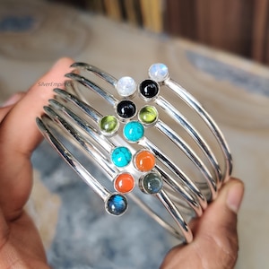Op de afbeelding: Een verzameling zilverkleurige armbanden, elk versierd met een gekleurde edelsteen. De edelstenen zijn onder andere turkoois, oranje, zwart en groen. De armbanden zijn op elkaar gestapeld, wat de verscheidenheid aan kleuren en ontwerpen laat zien. Afmetingen in centimeters.