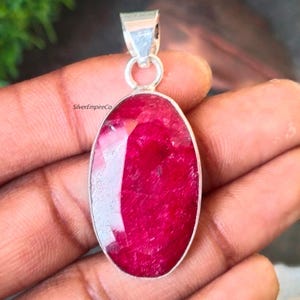 Pode incluir: Um pingente de prata com uma grande gema de rubi de forma oval. O rubi é de cor vermelho escuro e tem um corte facetado. O pingente está em uma corrente de prata.