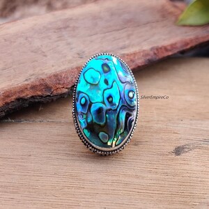 Könnte beinhalten: Ein silberner Ring mit einem ovalen Einlegeteil aus Abalone-Schale. Die Schale hat ein blaues, grünes und schwarzes irisierendes Muster. Der Ring ist in einer Silberfassung gefasst.