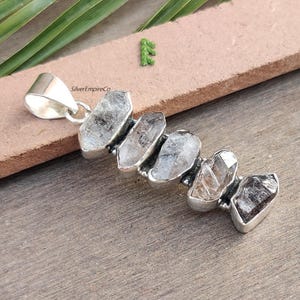 Pu&ograve; includere: Ciondolo in argento con un gruppo di cinque cristalli di quarzo trasparenti, ognuno con una forma e una dimensione uniche. I cristalli sono incastonati in argento e hanno un aspetto grezzo e naturale. SilverEmpireCo &egrave; stampato sul ciondolo.