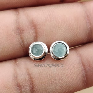 May include: A pair of silver stud earrings featuring light blue round gemstones. The earrings are set in a simple silver bezel setting.