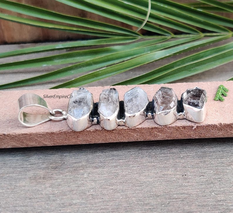 Pu&ograve; includere: Ciondolo in argento con cinque punte di cristallo di quarzo trasparente incastonate in argento, con un'anellina in argento. Il ciondolo &egrave; su una superficie marrone. SilverEmpireCo &egrave; visibile nell'immagine.