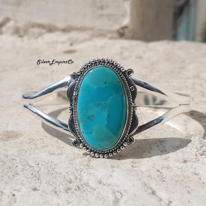 May include: A silver cuff bracelet with a large, oval turquoise stone. The turquoise is a vibrant blue-green colour, set in a decorative bezel. The bracelet has a simple, elegant design with curved arms.