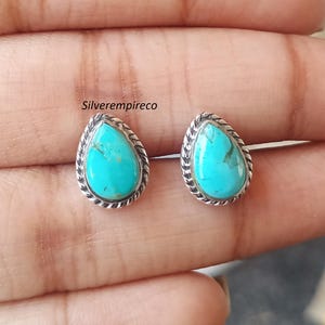 May include: A pair of turquoise teardrop stud earrings. Each earring has a vibrant blue-green stone set in a silver-toned, rope-textured bezel. The earrings are displayed against a neutral background.