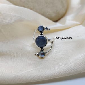 May include: A silver ring featuring three circular, blue-grey gemstones. The ring has an unusual, asymmetrical design with the text "Silver Empire Co" visible. The gemstones are different sizes, with the largest in the middle.