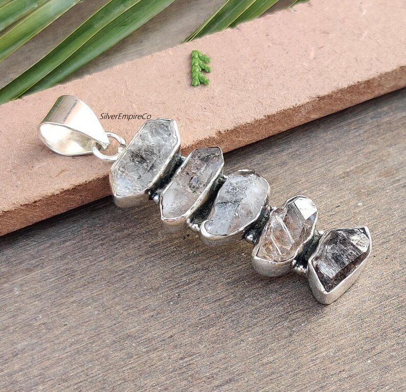 Pu&ograve; includere: Ciondolo in argento con un gruppo di cinque cristalli di quarzo trasparenti, ognuno con una forma e una dimensione uniche. I cristalli sono incastonati in argento e hanno un aspetto grezzo e naturale. SilverEmpireCo &egrave; stampato sul ciondolo.
