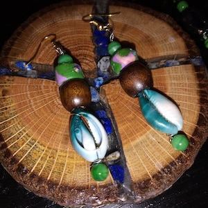 May include: A pair of dangle earrings featuring gold-tone hooks, green and purple patterned beads, dark brown wooden beads, and white and teal cowrie shells. Each earring is finished with a small green bead at the bottom, creating a layered design.