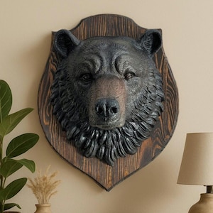 May include: A black bear head mounted on a wooden plaque with a dark wood grain. The bear has a realistic fur texture and a serious expression.