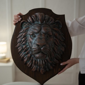 Pode incluir: Um escudo de madeira com uma cabeça de leão esculpida em relevo. A cabeça de leão é pintada em cobre com uma pátina verde. O escudo é segurado por uma mão.
