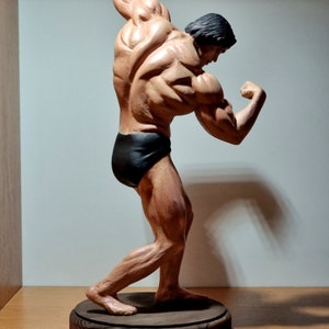 K&ouml;nnte beinhalten: Eine Skulptur einer muskul&ouml;sen Figur in einem schwarzen Posing-Anzug, die auf einem Holzsockel steht. Die Figur ist mit einem Arm &uuml;ber dem Kopf erhoben und dem anderen Arm im Ellbogen gebeugt.