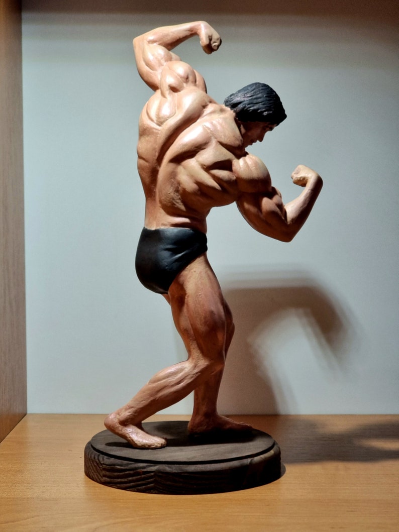 K&ouml;nnte beinhalten: Eine Skulptur einer muskul&ouml;sen Figur in einem schwarzen Posing-Anzug, die auf einem Holzsockel steht. Die Figur ist mit einem Arm &uuml;ber dem Kopf erhoben und dem anderen Arm im Ellbogen gebeugt.