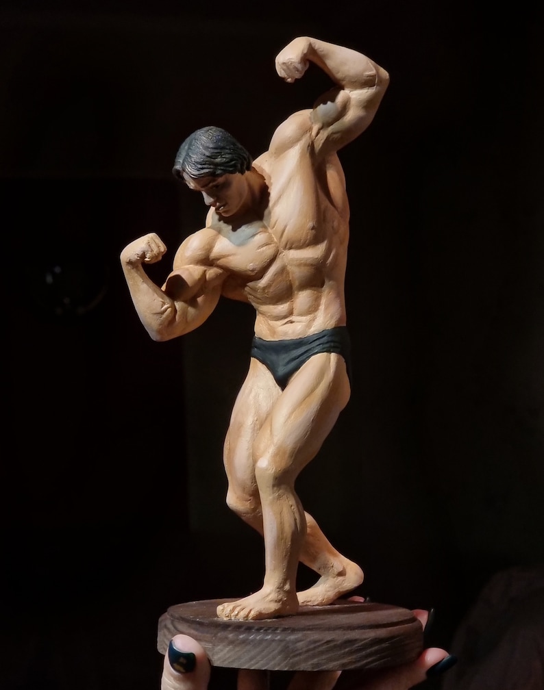 K&ouml;nnte beinhalten: Eine Skulptur eines muskul&ouml;sen Mannes in einem schwarzen Badeanzug, der mit gebeugten Armen posiert. Die Figur steht auf einem Holzsockel.