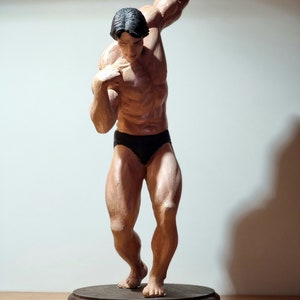 K&ouml;nnte beinhalten: Eine Skulptur eines muskul&ouml;sen Mannes in einem schwarzen Badeanzug, die auf einem Holzsockel steht. Die Figur ist mit einem erhobenen Arm und dem anderen Arm, der im Ellbogen gebeugt ist, dargestellt.