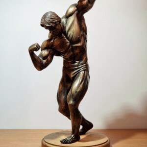K&ouml;nnte beinhalten: Eine bronzene Figur eines muskul&ouml;sen Mannes, der mit seinen Armen posiert. Die Figur steht auf einem Holzsockel.