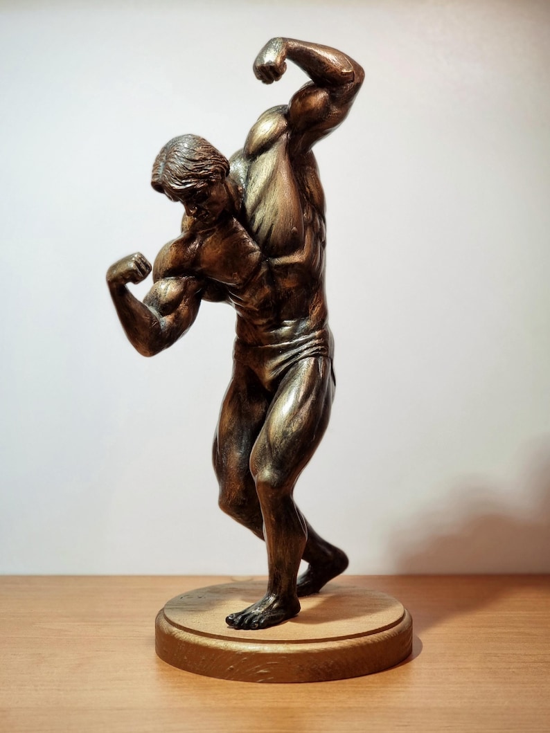 K&ouml;nnte beinhalten: Eine bronzene Figur eines muskul&ouml;sen Mannes, der mit seinen Armen posiert. Die Figur steht auf einem Holzsockel.