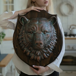 Könnte beinhalten: Eine Holzplakette mit einem geschnitzten kupferfarbenen Bärenkopf. Der Bär hat ein strukturiertes Fell und einen realistischen Ausdruck. Die Plakette ist dunkelbraun mit einer Holzmaserung.