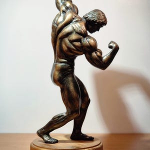 K&ouml;nnte beinhalten: Eine bronzene Skulptur einer muskul&ouml;sen Figur in einer Bodybuilding-Pose. Die Figur steht auf einem Holzsockel.