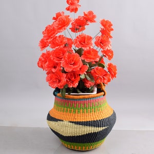 May include: A colourful woven basket with a black, orange, green and white pattern. The basket is filled with a bouquet of bright orange silk flowers.
