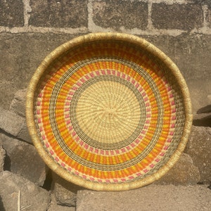 May include: A round woven basket with a natural straw base and colorful stripes in shades of orange, pink, and yellow.