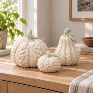 Puede incluir: Un conjunto de tres calabazas decorativas de color blanco roto con un patrón de punto de ochos texturizado. Cada calabaza está rematada con un tallo de color verde salvia claro y un pequeño detalle de hoja. Las calabazas varían en tamaño y forma, incluyendo una redonda, una pequeña y una alargada.