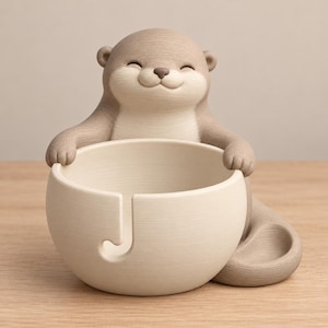 May include: A light brown, smiling otter figurine holding a cream-colored yarn bowl. The bowl features a smooth, rounded shape with a J-shaped cutout on the front for yarn to pass through. The otter has a textured surface and a happy expression.