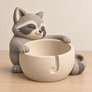 Puede incluir: Una figura de mapache gris y blanco con los ojos cerrados, apoyando sus patas sobre un cuenco para lana de color crema. El cuenco presenta un diseño liso y redondeado con un corte en forma de J en la parte frontal para guiar el hilo y cola.