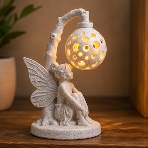 Peut inclure: Une figurine en résine blanc cassé mouchetée représentant une fée assise avec des ailes, placée sous une structure en branche courbée qui soutient une lanterne sphérique perforée diffusant une lumière chaude par ses ouvertures.