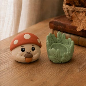 May include: A clay figurine of a mushroom character with a red cap and white spots, holding a small brown acorn, positioned next to a separate green clay holder shaped like a cluster of leaves with a central circular opening for holding items.