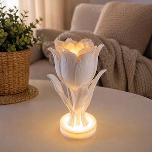Può includere: Una lampada bianca a forma di tulipano con petali e foglie testurizzati, che emana una luce calda e luminosa dal centro del fiore e dalla base circolare. La lampada presenta una finitura opaca e poggia su una superficie liscia.