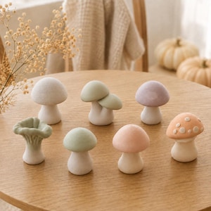 Puede incluir: Una colección de siete pequeñas figuras de setas en varios colores pastel, incluyendo blanco, verde salvia, lavanda, melocotón y rosa. Las setas presentan sombreros y tallos lisos, con una seta verde de bordes ondulados y una seta con puntos.