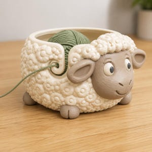 Op de afbeelding: Een crèmekleurige keramische garenkom in de vorm van een schaap met een textuur van wol, een glad bruin gezicht en vier kleine bruine voetjes. Een groene bol garen ligt in de kom, waarbij de draad door een spiraalvormige opening naar buiten komt.