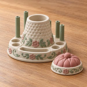 May include: A cream-colored sewing organizer featuring a central thimble-shaped holder, four vertical green posts, and small circular slots. The base is decorated with pink floral motifs and green leaves, accompanied by a separate pink pincushion.