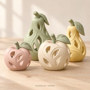 Puede incluir: Un conjunto de cuatro esculturas decorativas con forma de fruta y patrones de hojas recortadas. El set incluye una manzana rosa, una manzana crema, una pera verde salvia y una pera amarilla, cada una con un tallo y hoja verde.