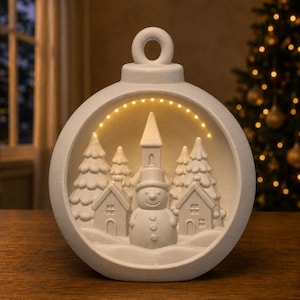 May include: A matte white, circular, hollow ornament featuring a 3D winter scene inside. The scene includes a central snowman, two small houses, and several pine trees. A string of warm yellow LED lights arches over the top of the scene.