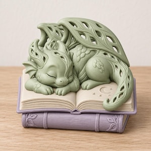 May include: A sage green dragon figurine with intricate cut-out wing details sleeping on an open book. The book rests on a closed lavender-colored book with embossed leaf patterns. The open page features a small crescent moon and star engraving.