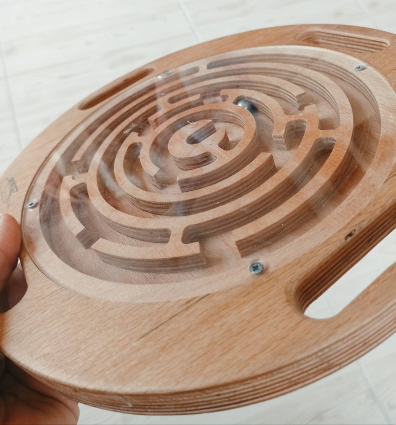 May include: A wooden circular maze with a clear plastic cover and a small metal ball inside. The maze has a complex pattern of grooves and channels.