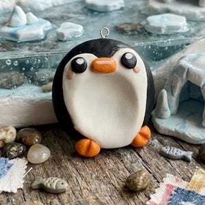 Può includere: Un ciondolo a forma di pinguino scolpito e rotondo con dorso nero, pancia bianca e becco e zampe arancioni. Il pinguino ha grandi occhi neri con riflessi bianchi e piccole guance rosa. Un piccolo anello in metallo argentato è fissato sulla parte superiore.