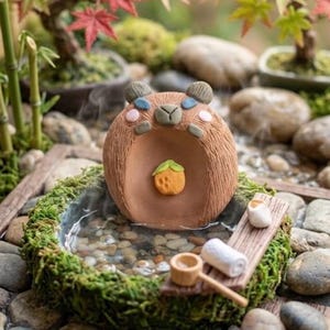 Può includere: Una statuetta in ceramica marrone a forma di capibara è posizionata in una vaschetta ricoperta di muschio, piena d'acqua e ciottoli. Il capibara ha un piccolo frutto arancione sul petto. Un asse di legno sostiene un asciugamano e un mestolo.