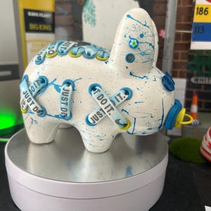 May include: White ceramic bunny figurine with blue and yellow accents. The bunny is decorated with blue paint splatters and has the words "Just Do It" written on its body.