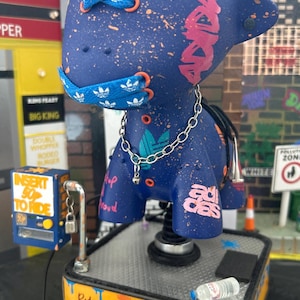 May include: A blue and pink toy horse with a silver chain and a blue and white Adidas logo on the chest. The horse is on a yellow base with the words "Ride Your Adidas Off".