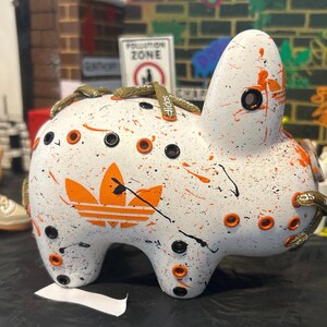 May include: White ceramic bunny figurine with orange and black paint splatters. The bunny has a large orange Adidas logo on its side and a green and white shoelace tied around its neck.