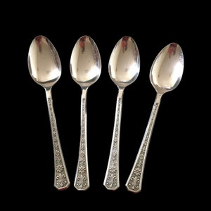 May include: Four silver spoons with oval bowls and ornate floral and fleur-de-lis designs on the handles. The spoons are arranged against a black background, showcasing their reflective surfaces and intricate details. A classic silverware set.