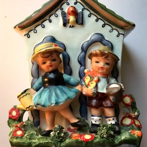 May include: Vintage ceramic wall pocket featuring two children in a garden scene. The girl wears a blue dress and yellow hat, holding a watering can. The boy holds flowers and a jug. A bird sits atop the roof.