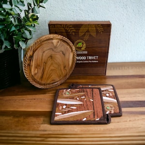 Pode incluir: Um descanso de panela redondo de madeira com acabamento em madeira natural. O descanso de panela é embalado em uma caixa com o texto "Acacia Wood Wood Trivet Organic Cotton Pot Holders". Também estão incluídos dois descansos de panela de algodão marrom e bege com um padrão de folhas.