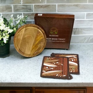Puede incluir: Un posavasos redondo de madera de mango con un juego de dos agarraderas de olla con estampado marrón y blanco. La caja dice "K&R 100% Madera de Mango Posavasos de Madera Premium Juego de Agarraderas de Olla de Algodón Orgánico 100%."