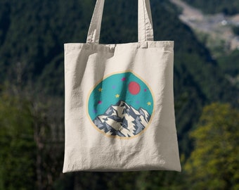 以下が含まれることがあります： クリーム色のトートバッグ。山脈、赤い円、黄色の星がターコイズブルーの背景に描かれたデザイン。バッグは緑の山々を背景に掲げられています。