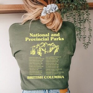 May include: Olive green t-shirt with the text "National and Provincial Parks" and "British Columbia" in yellow. A mountain graphic is centered between the text. The person is wearing blue jeans.