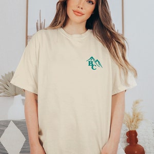 May include: Cream-colored t-shirt with a teal mountain graphic and the letters "BC". The shirt has short sleeves and a relaxed fit. The model is wearing neutral-colored shorts.